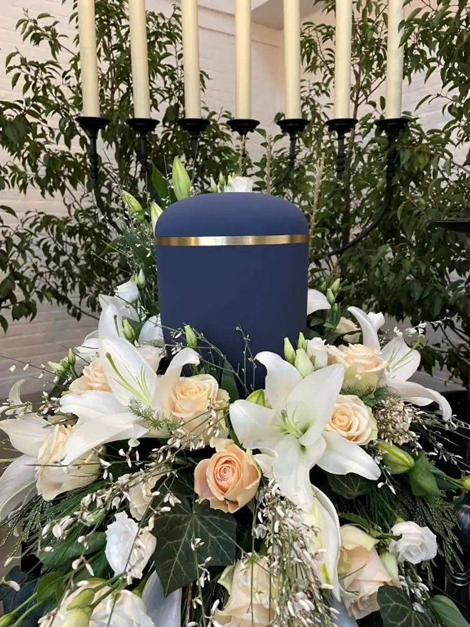 Urne mit weißem Lilien- und Rosenschmuck vor einem Kerzenleuchter