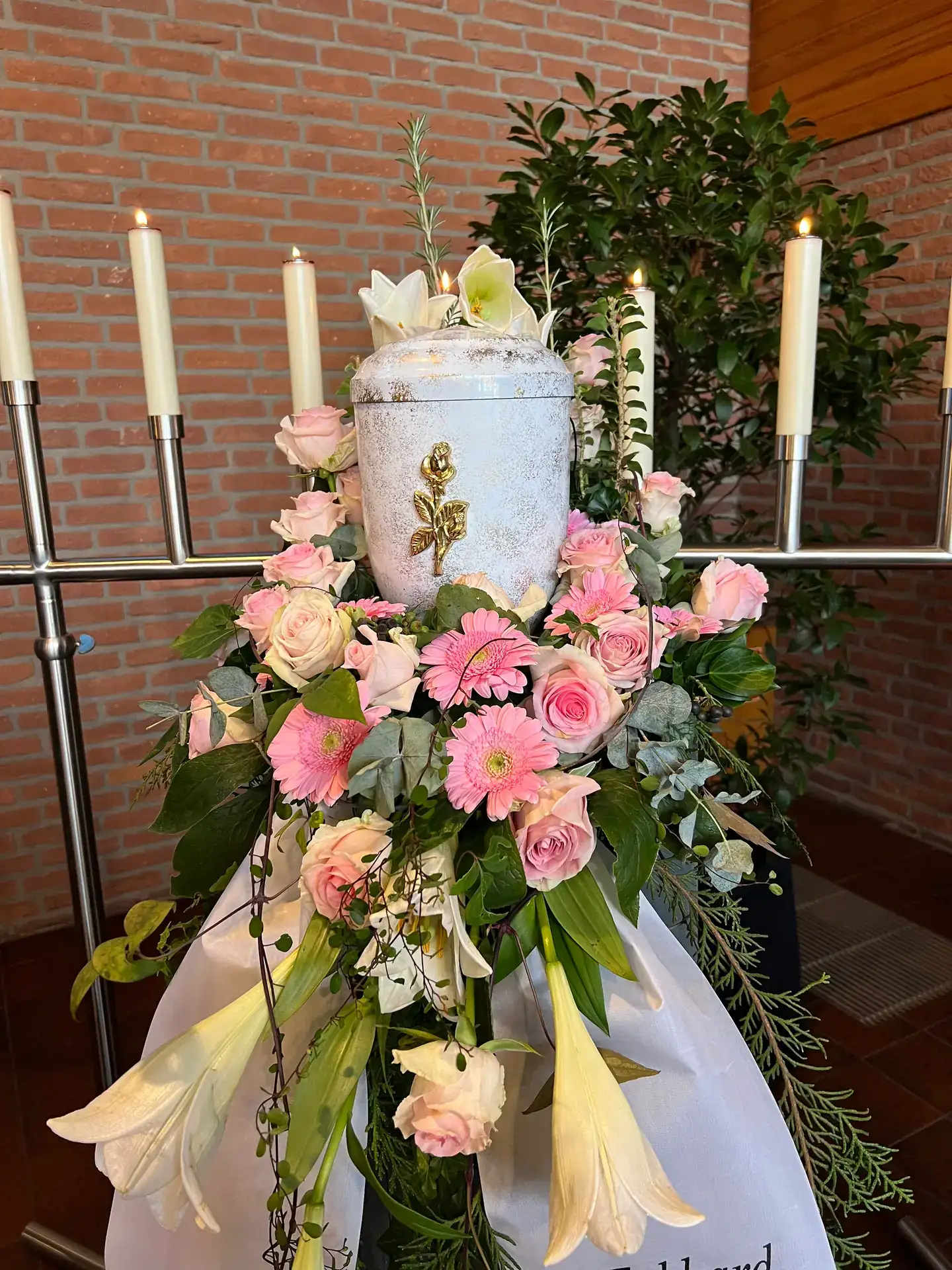 Helle Urne mit goldener Rose, rosa Gerbera und Rosen zwischen Kerzenleuchtern