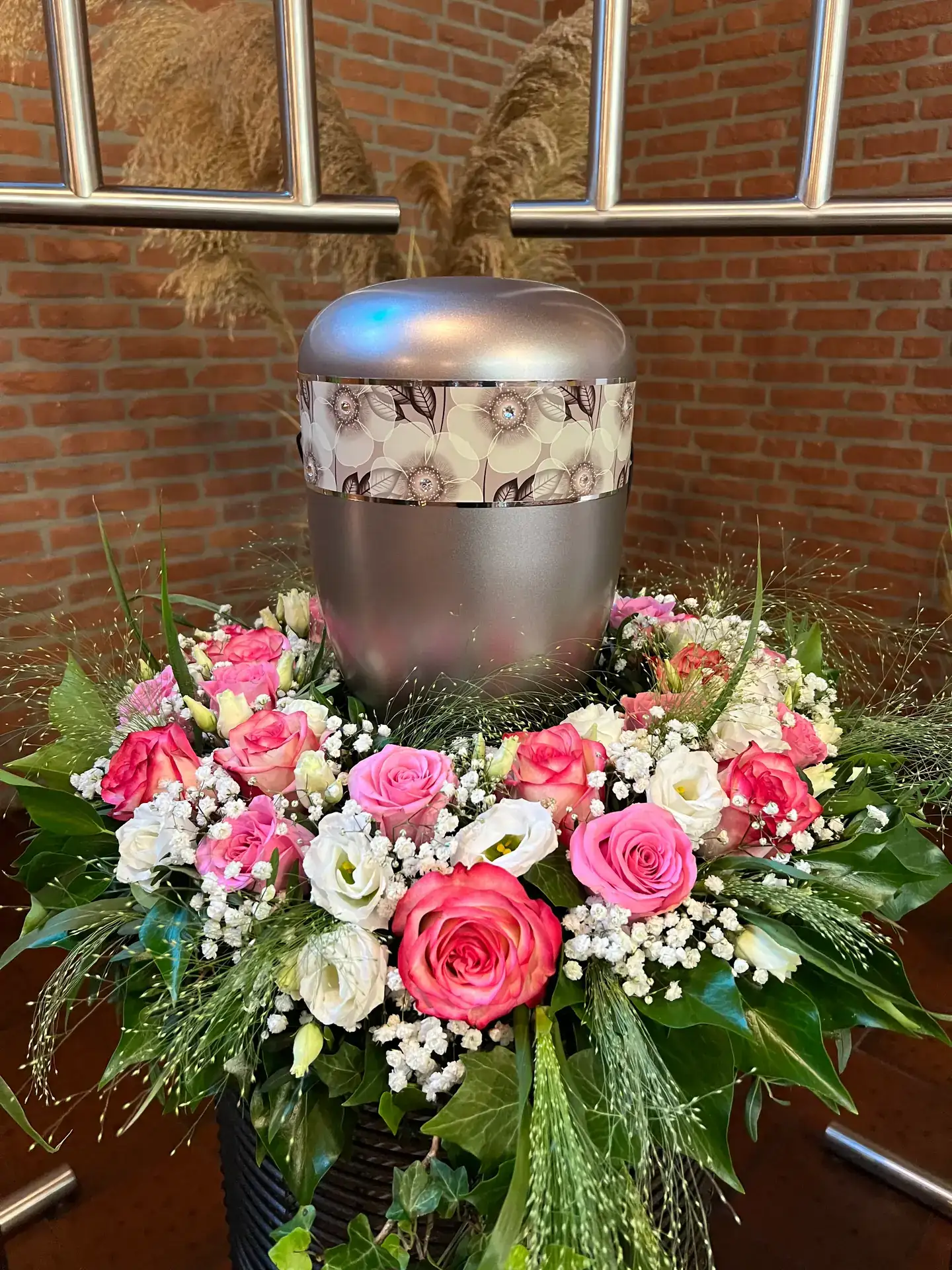 Urne mit Blütenband und einem Kranz aus Rosen vor einer Backsteinwand
