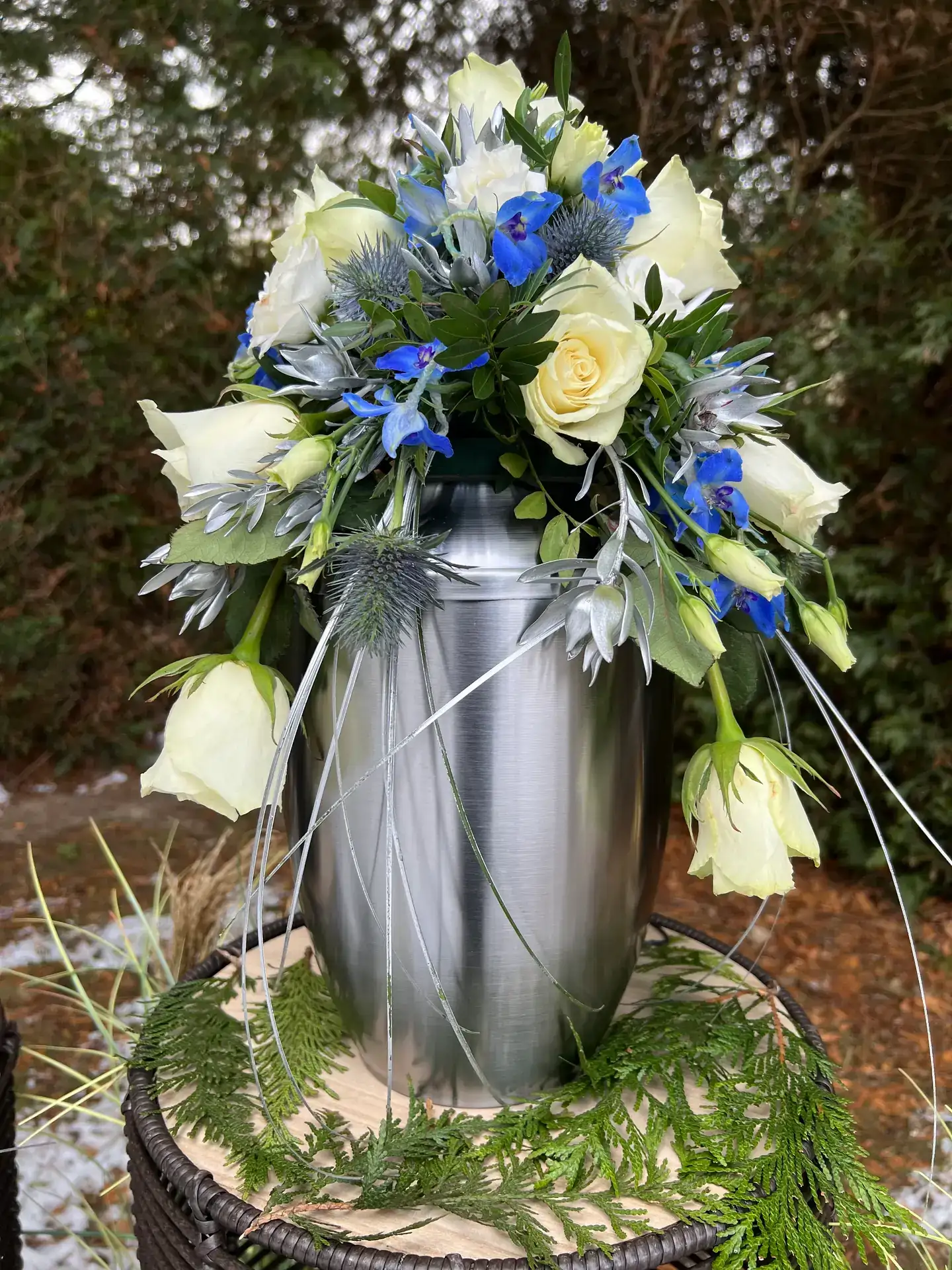 Silberne Urne mit blau-weißem Blumenschmuck im Freien auf einem Baumstumpf